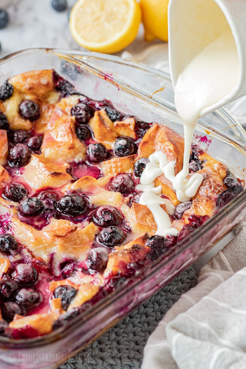 Baked lemon blueberry bread pudding in glass pan with lemon icing pouring on top. 