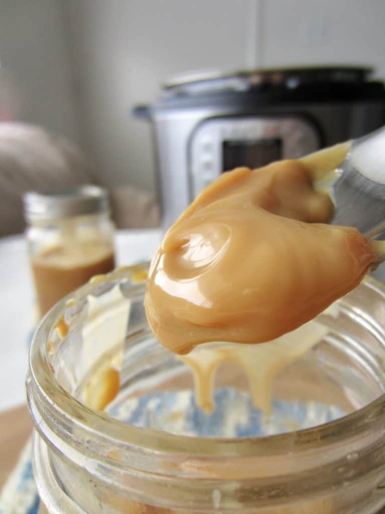 A spoon above a jar with dulce de leche, instant pot in the background. 