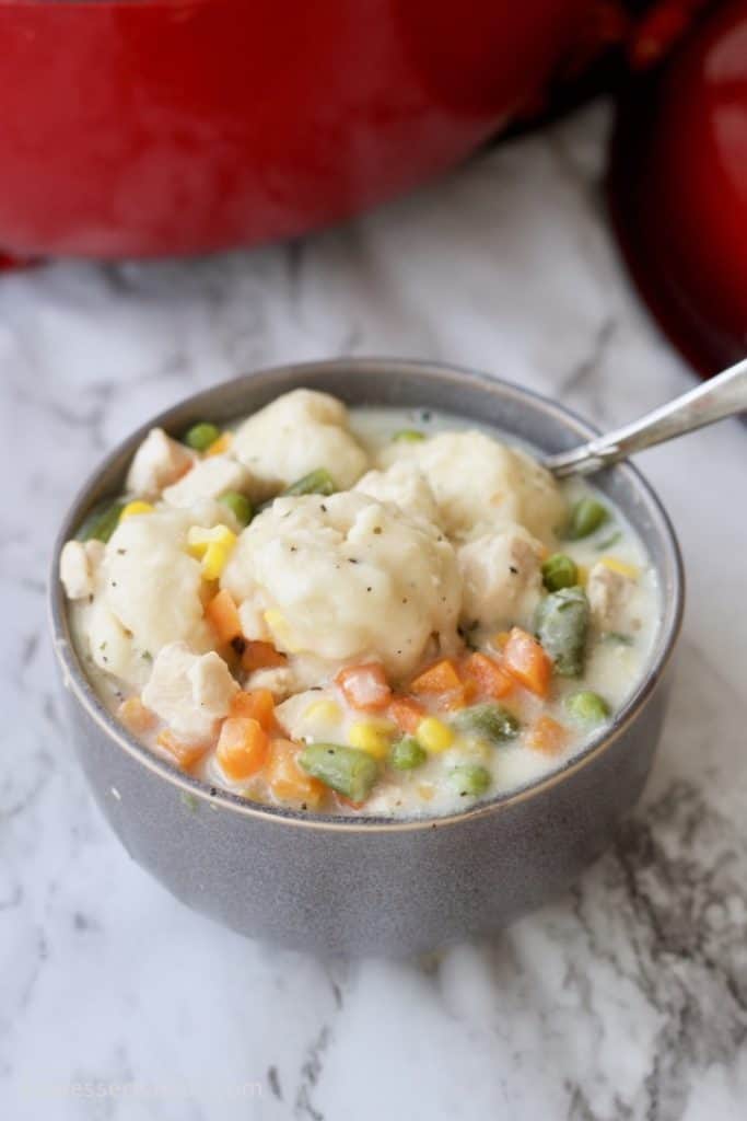 Grey bowl full of creamy chicken and dumplings, carrots, corn, peas and green beans with a spoon on top of a marble countertop. 