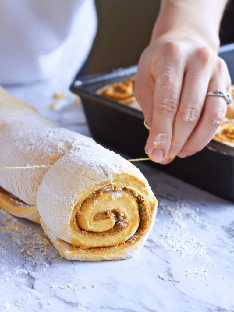 Pumpkin Cinnamon Roll raw, being cut with string to place in pan on a floured marble surface. 
