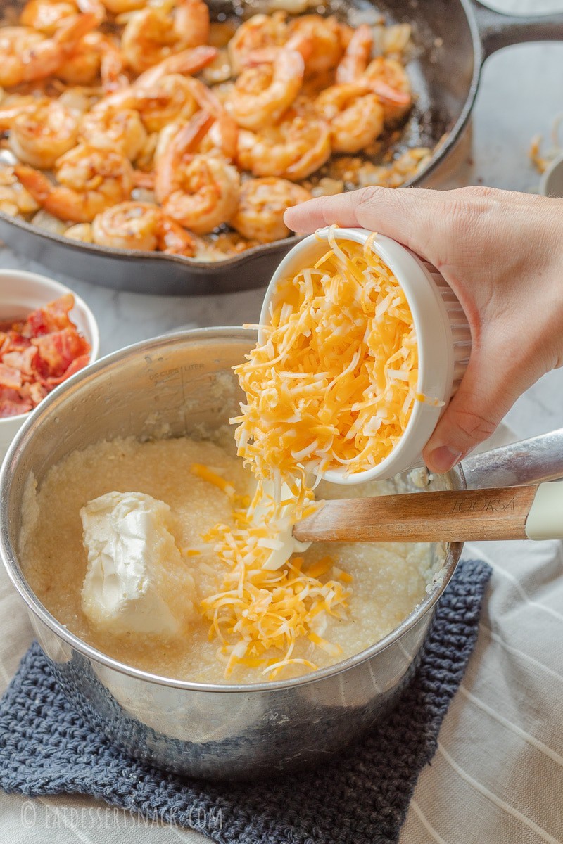Pouring dish of shredded cheddar cheese into pan of cooked grits and cream cheese. 