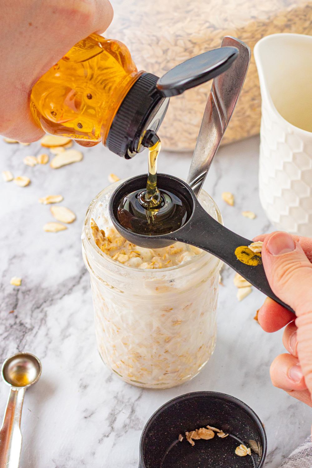 honey in tablespoon over oats in a mason jar with white cup and oats in background. 