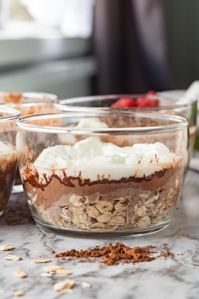 clear glass bowl with layers of oats, cocoa powder, yogurt, and milk. 