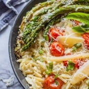 Black pan of cooked orzo, roasted asparagus and tomatoes, parmesan cheese on top with blue tea towel.