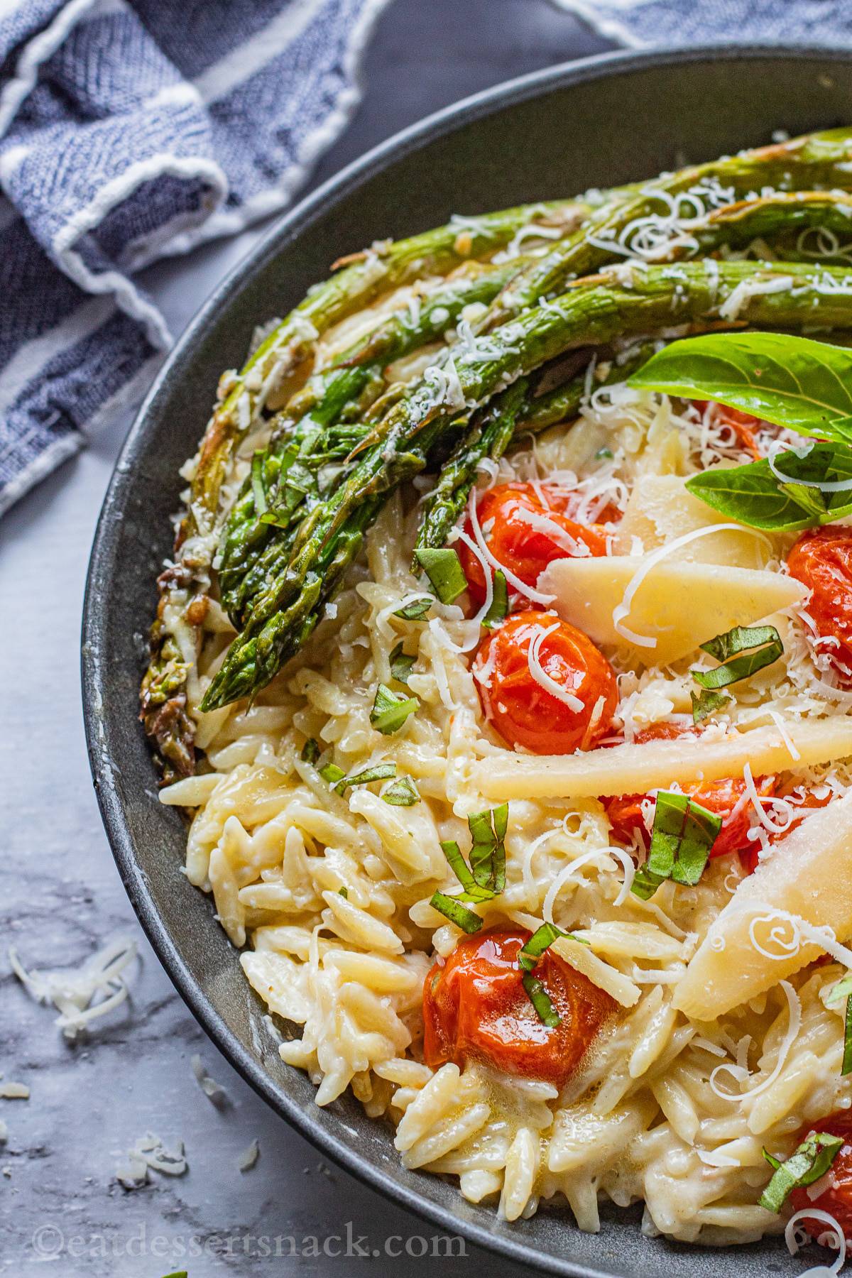 Black pan of cooked orzo, roasted asparagus and tomatoes, parmesan cheese on top with blue tea towel.