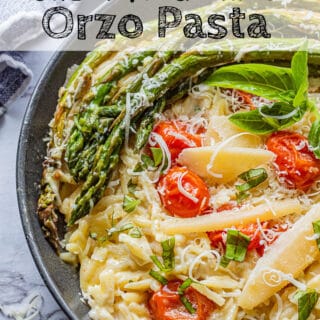 cooked orzo pasta in black pan with roasted asparagus and tomatoes on top with parmesan cheese and basil leaves. Text overlay reads creamy garlic orzo pasta.
