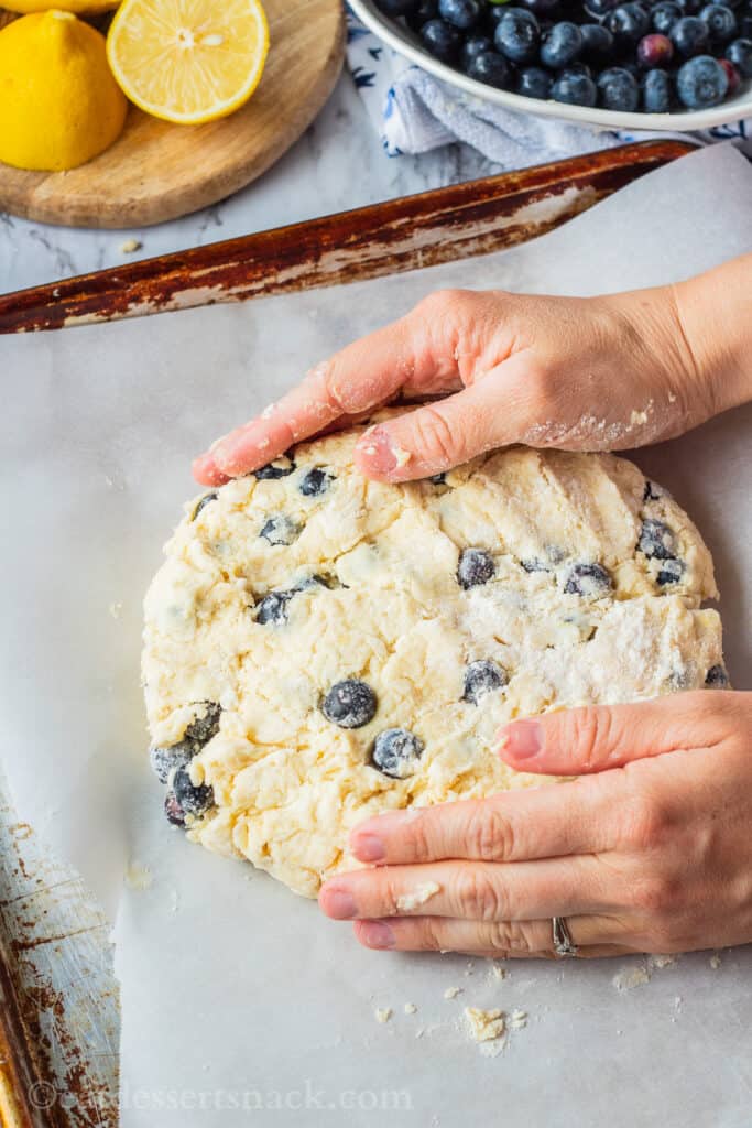 hands patting circle of raw blueberry scone dough onto metal pan. 