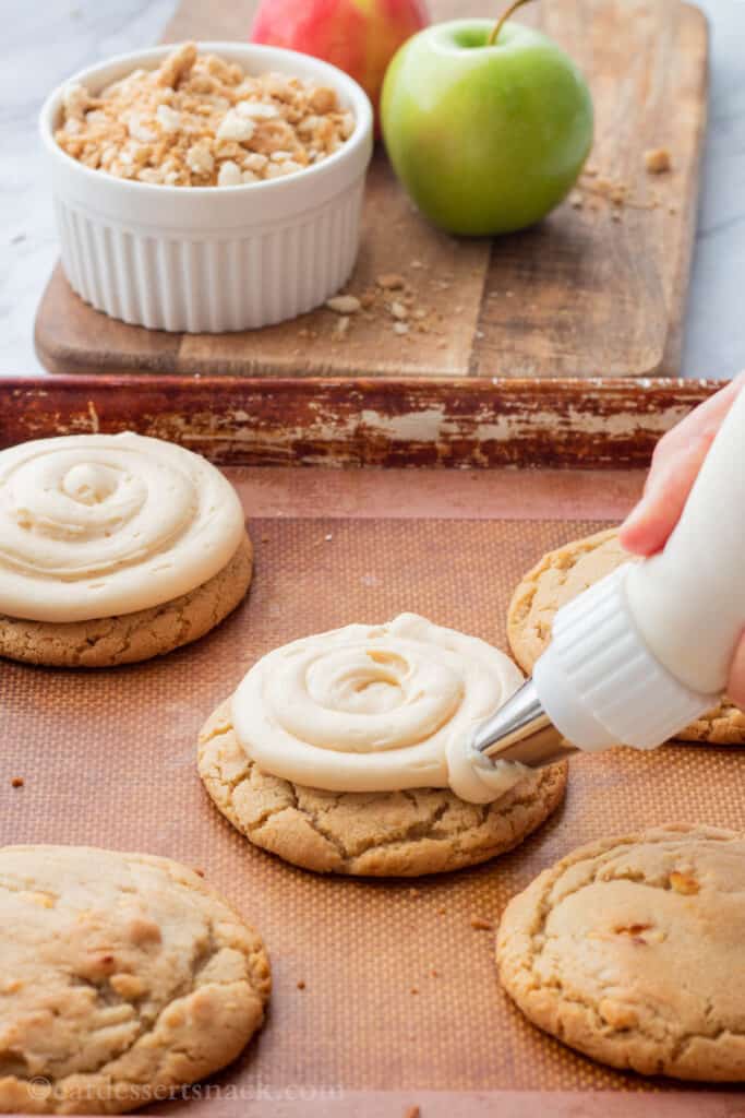 Piping frosting onto baked caramel apple cookies on baking sheet. 