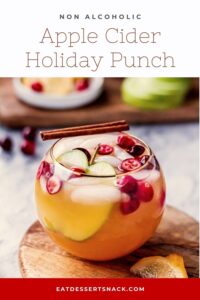 Apple cider punch in round glass with cranberries and cinnamon stick on wood board.