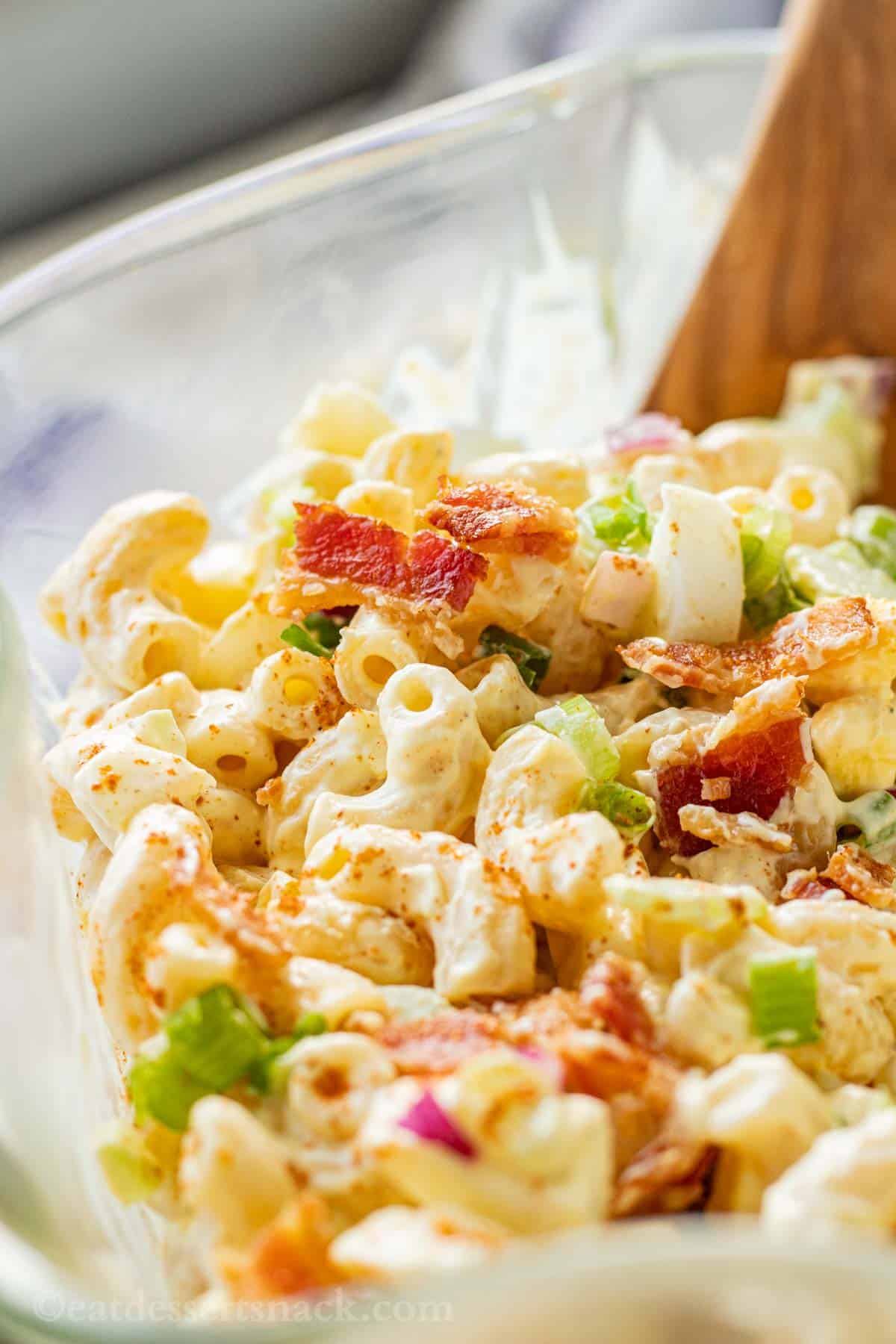 Deviled egg pasta salad in clear glass bowl with wooden spoon and bacon on top. 