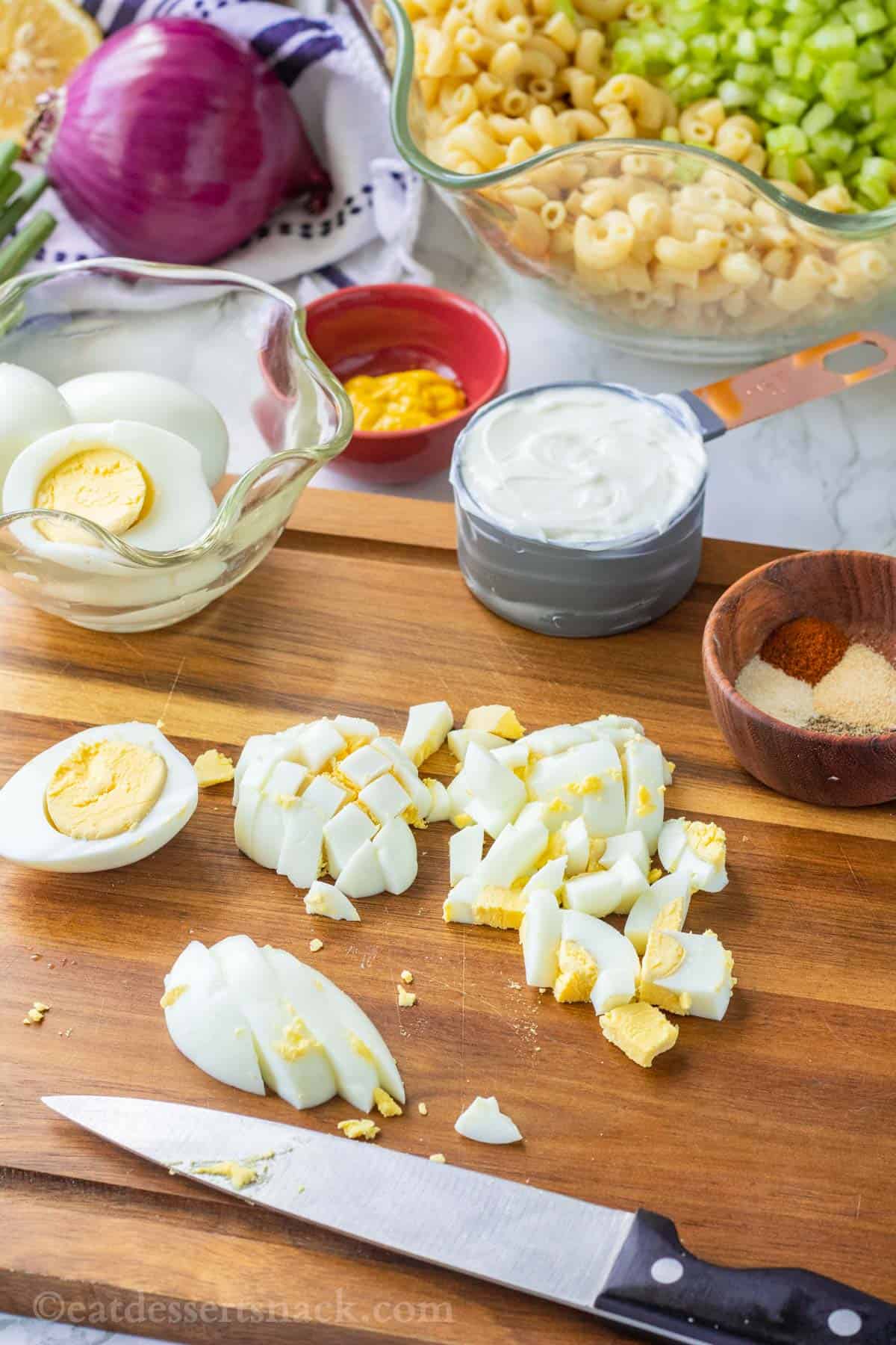 Knife with hard boiled eggs being chopped on wood cutting board with spices and mayo in background.