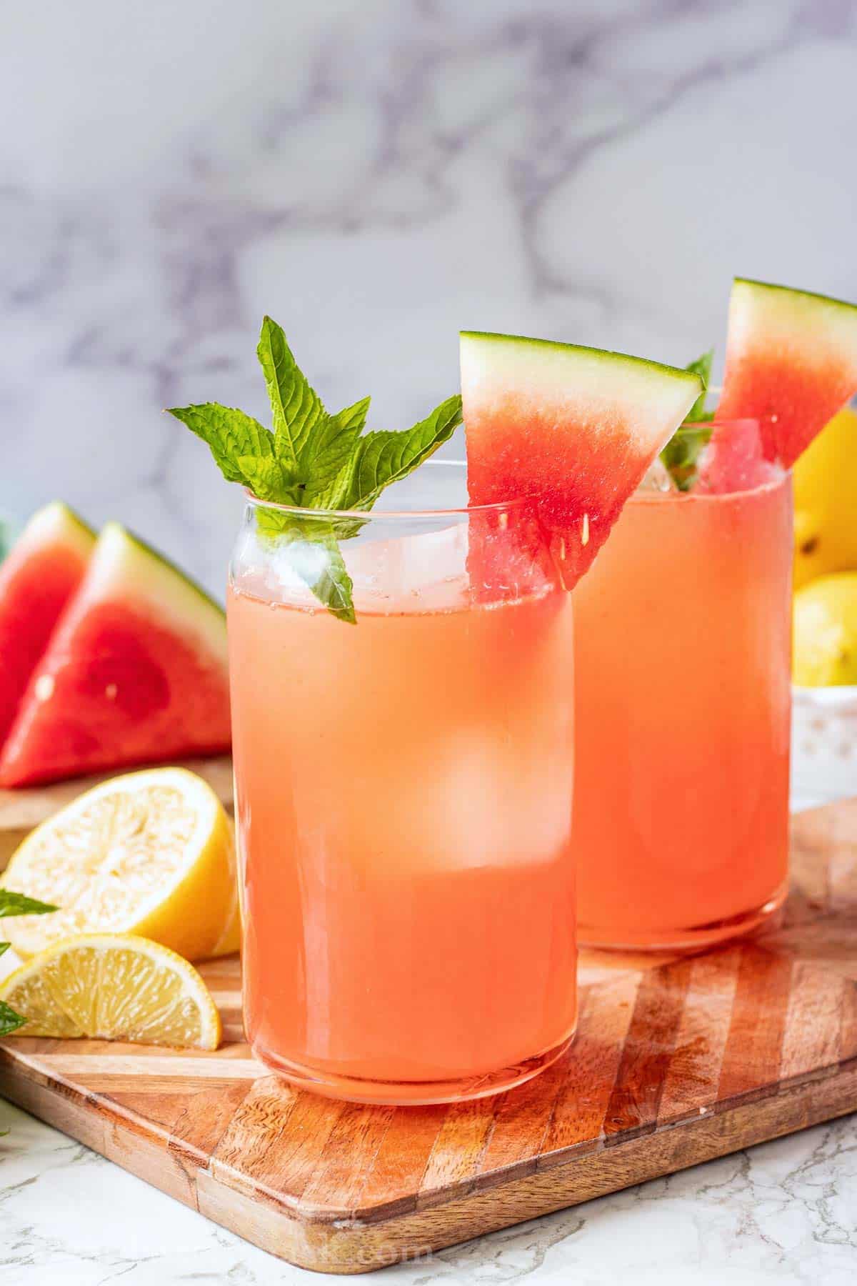 2 glasses of watermelon lemonade with watermelon wedge on rim and mint sprig on wood cutting board.