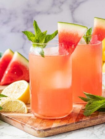 2 glasses of watermelon lemonade with sprigs of mint and watermelon wedges on a wood cutting board.