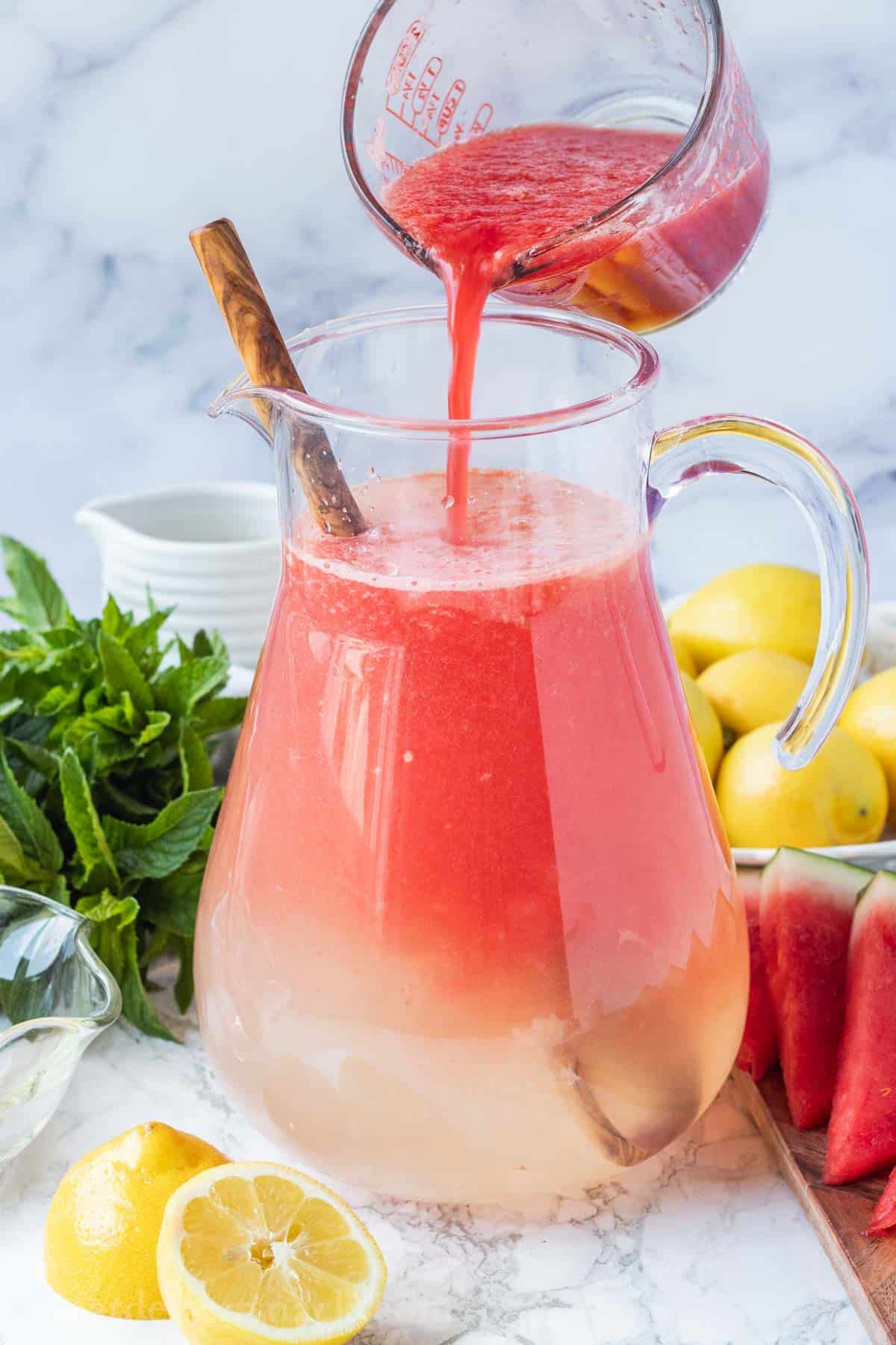 Pouring watermelon juice into clear pitcher of water with mint, lemons, and watermelon in background.