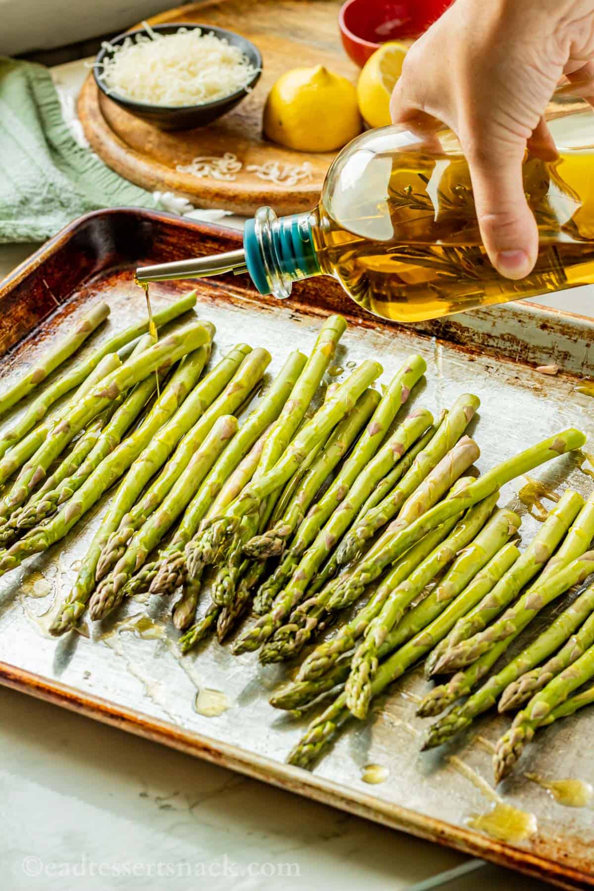 Hand sprinkling bottle of olive oil over metal pan fo raw asparagus.