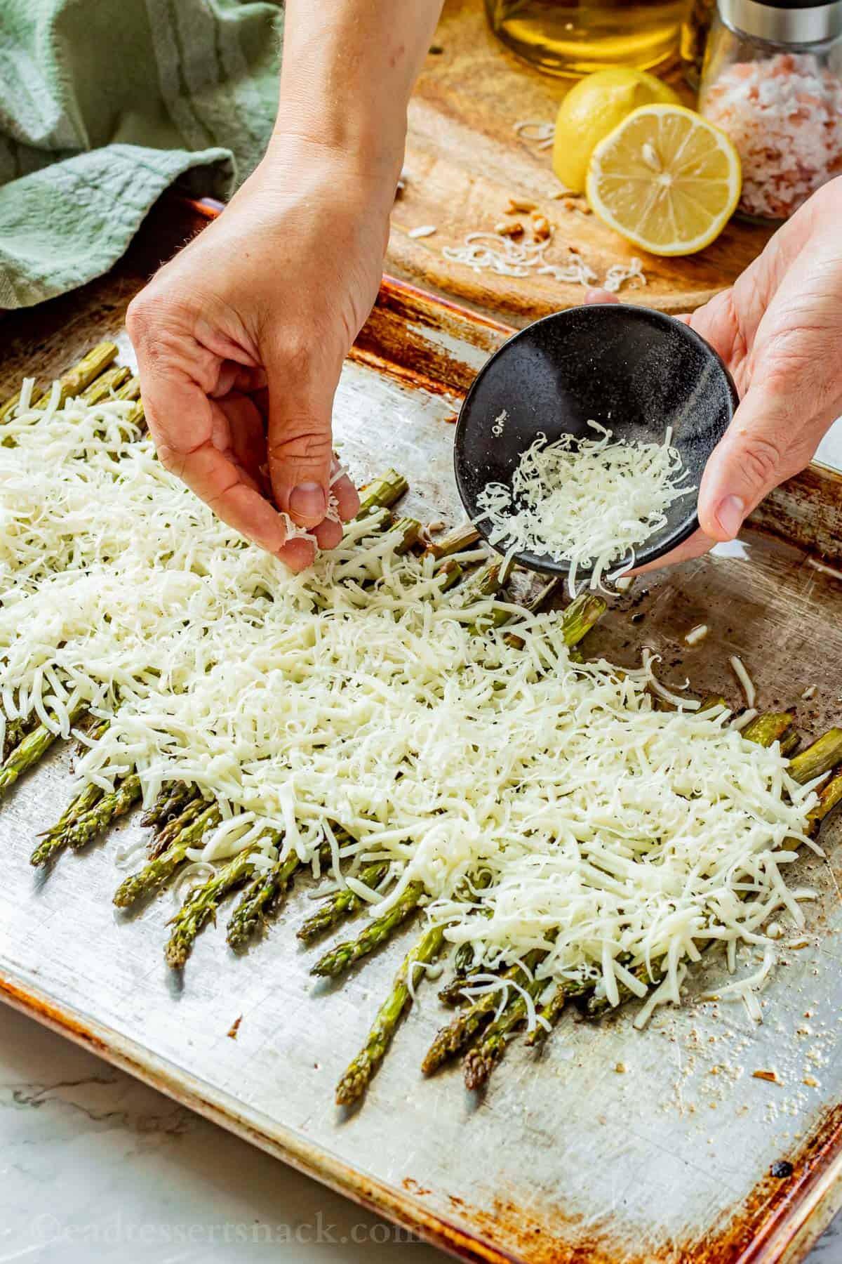 Hand sprinkling cheese over baked asparagus on metal pan.