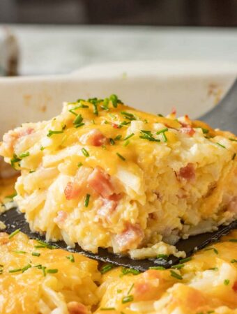 Slice of baked ham and cheese hash brown casserole on black spatula above white casserole dish.