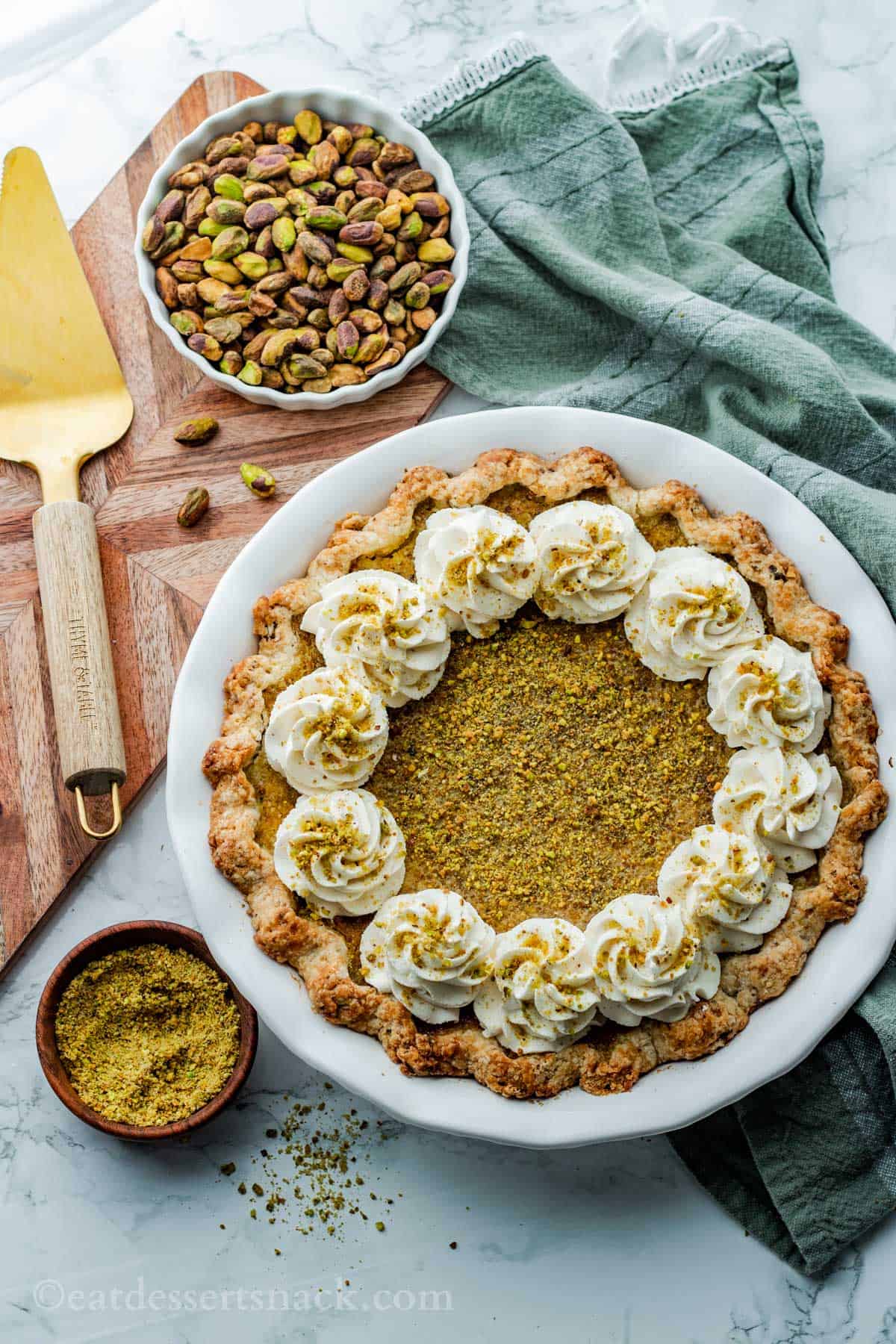 Baked pistachio pie with spatula and bowl of pistachios with green dish towel on marble countertop.