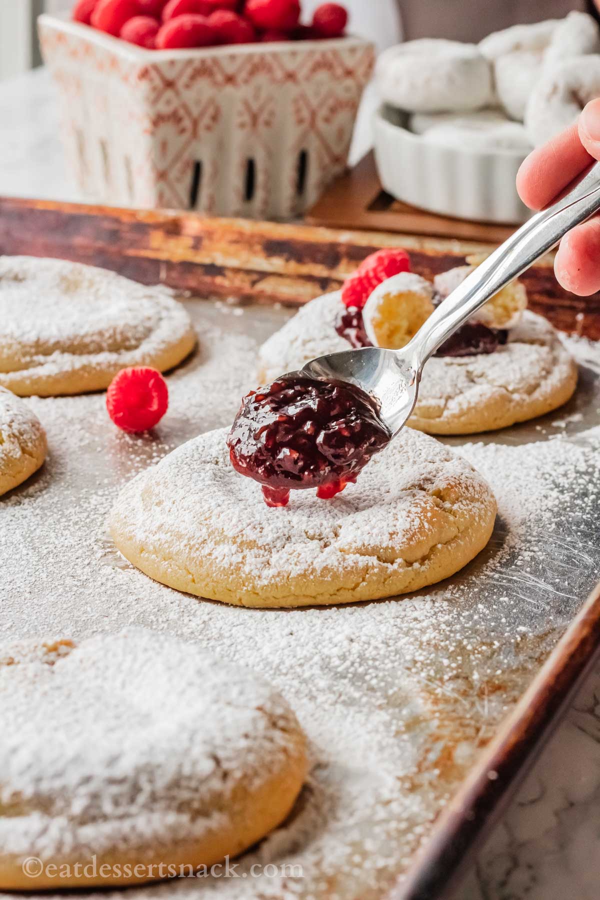 Metal spoon with raspberry jam above baked vanilla cookie covered in powdered sugar, on metal pan.