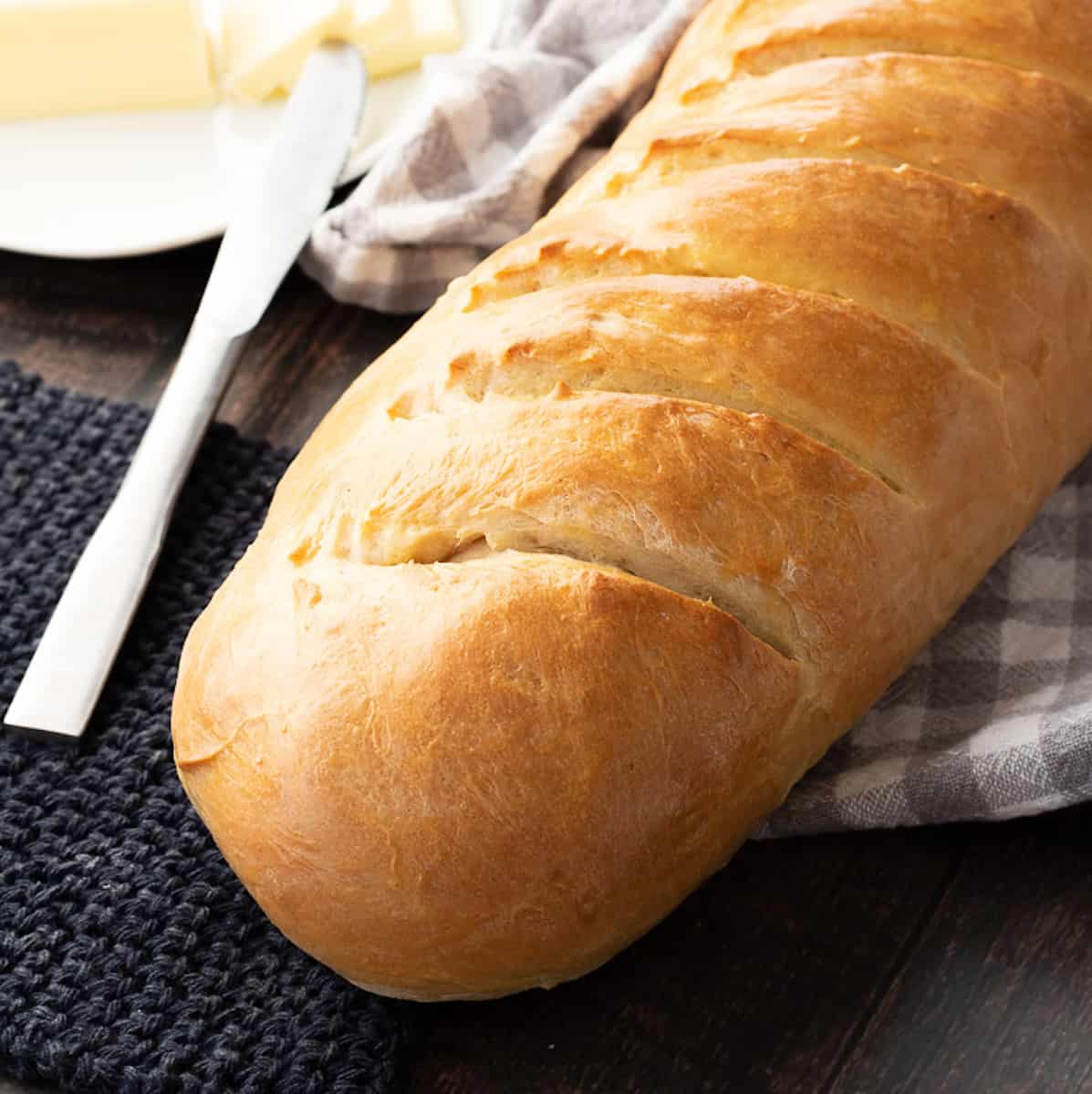 Baked loaf of fluffy french bread on wood table with towel, butter, and knife.