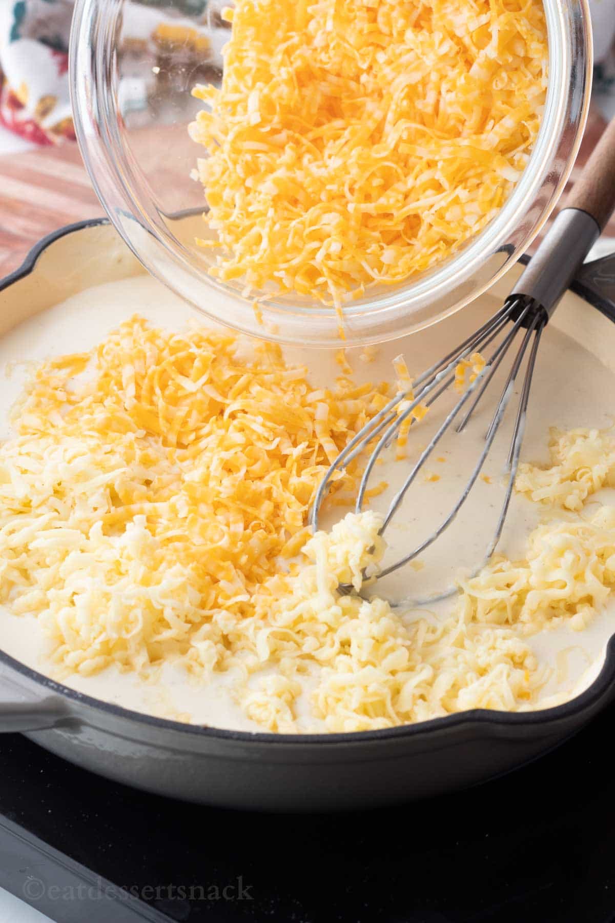 Glass bowl of cheese pouring into frying pan of milk sauce with wire whisk.