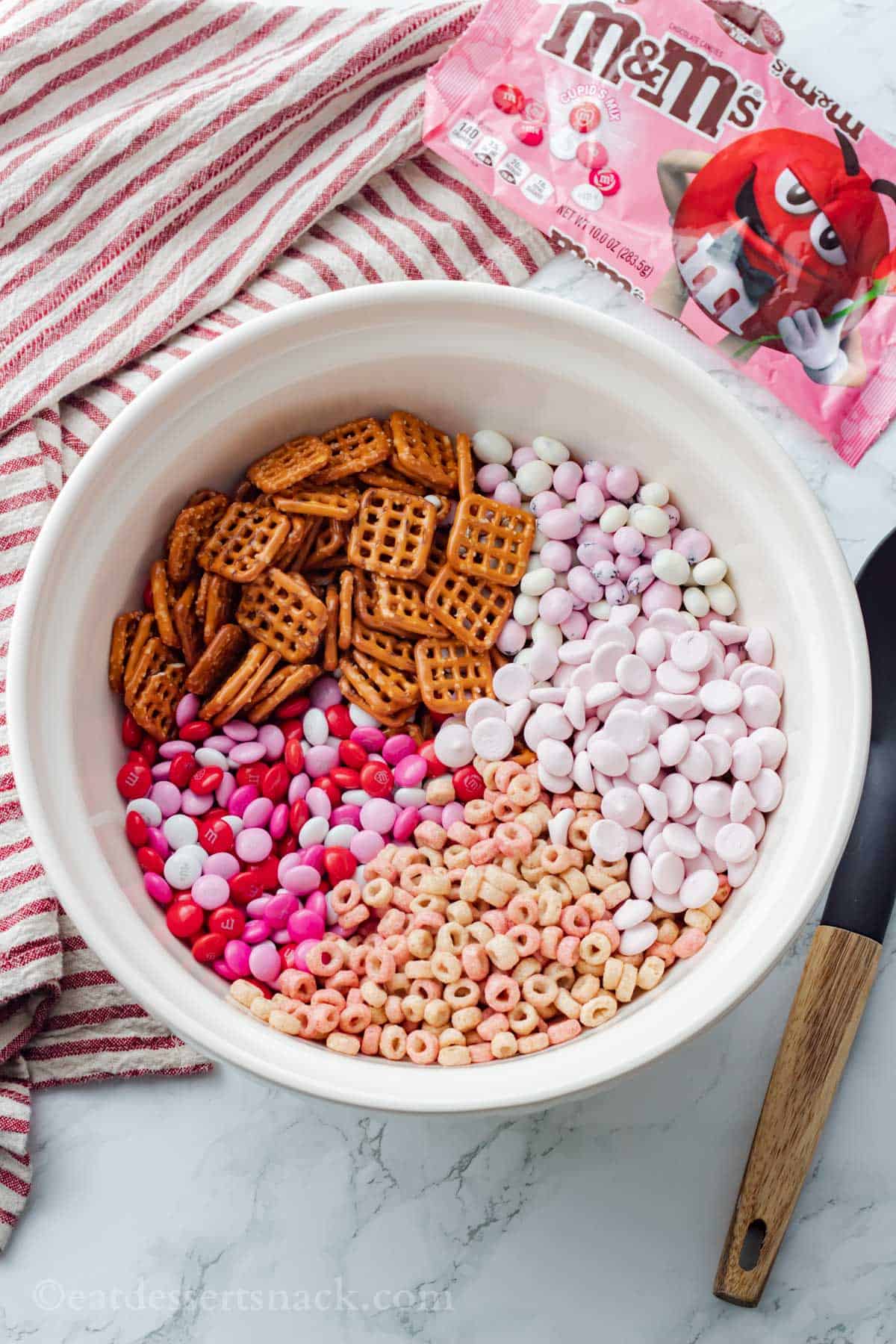 Ingredients for Valentines Trail Mix in a large white ceramic bowl on marble with wooden spoon and red and white striped towel.