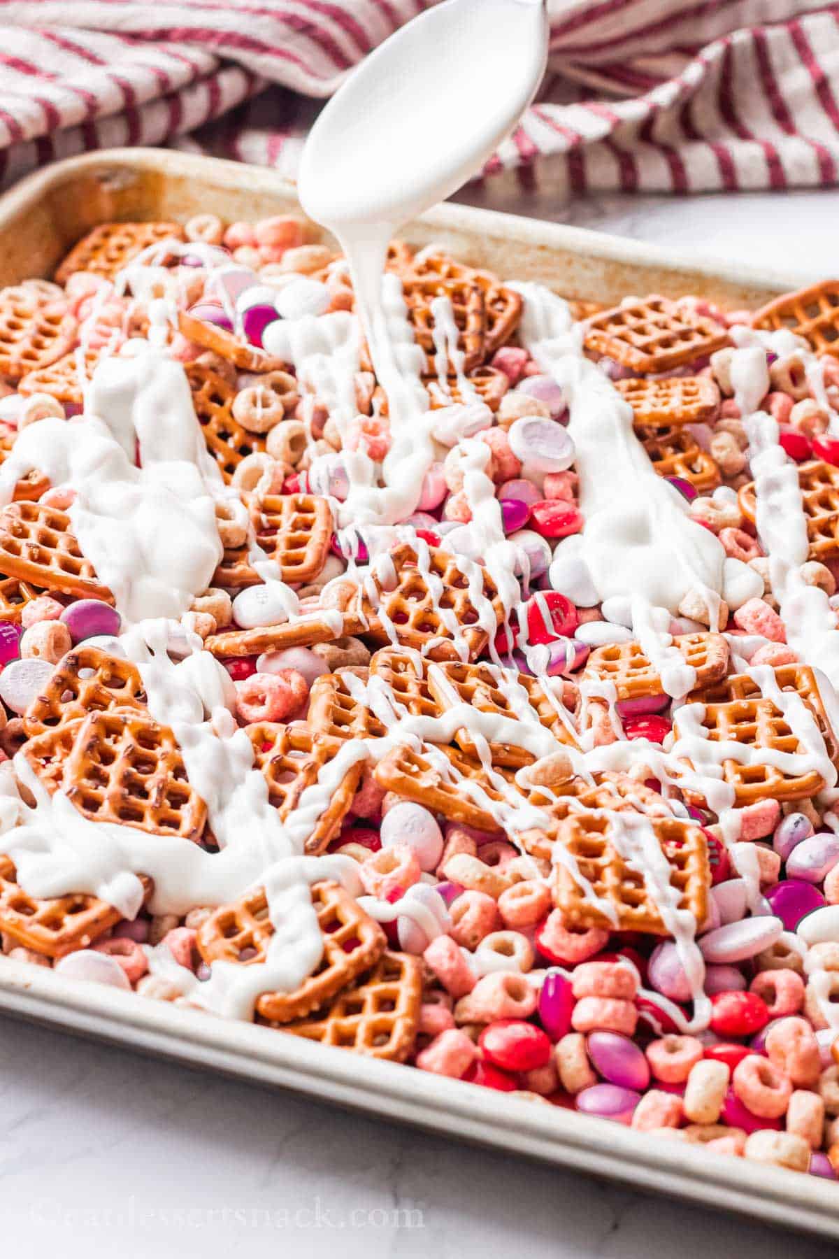 Pretzels, pink and red M&Ms, Cheerios, yogurt melts, and yogurt covered raisins on a metal baking sheet with white chocolate drizzling over them on a spoon.
