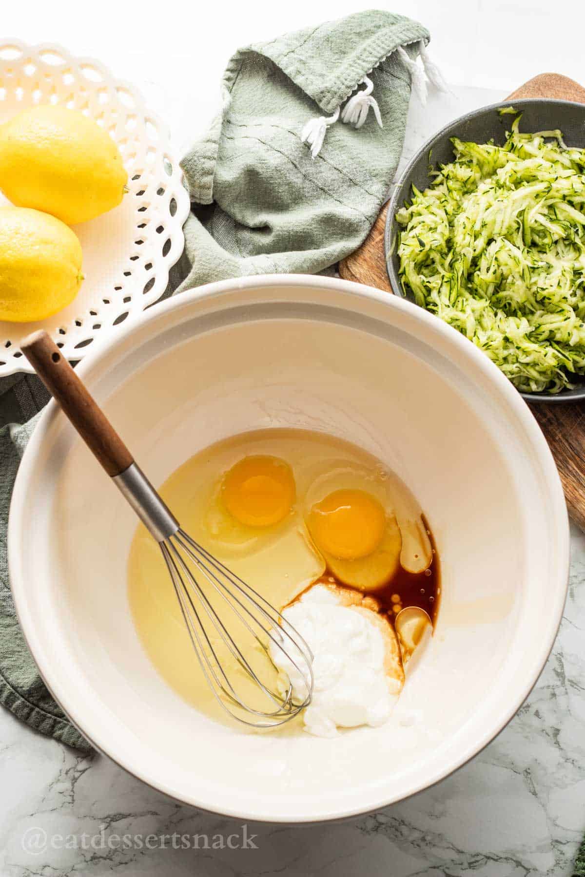 Eggs, greek yogurt, oil, and vanilla in ceramic mixing bowl with wire whisk.