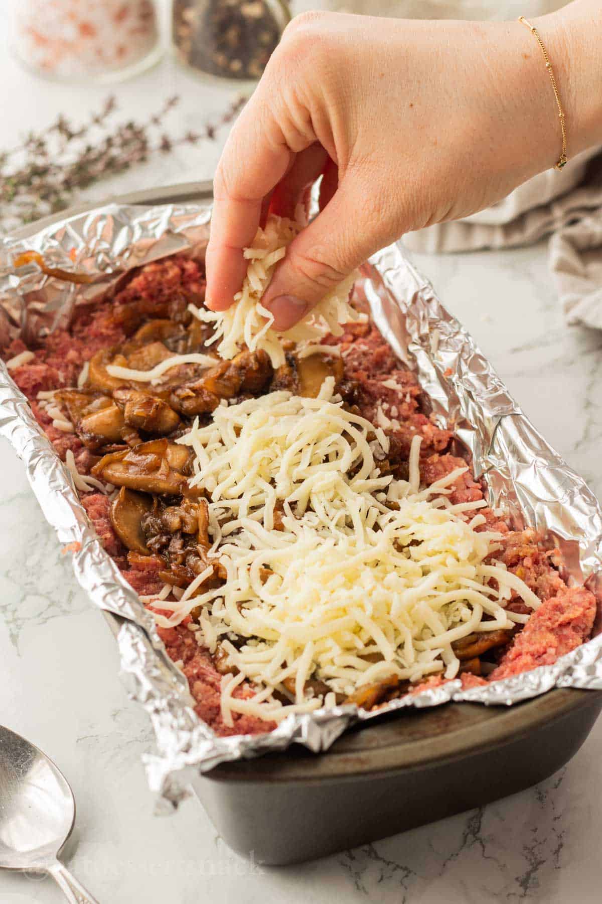 Hand sprinkling cheese onto cooked onions and mushrooms in raw beef in metal pan.