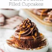 Unwrapped chocolate caramel cupcake frosted, sitting on white dish on marble countertop with pan of cupcakes.
