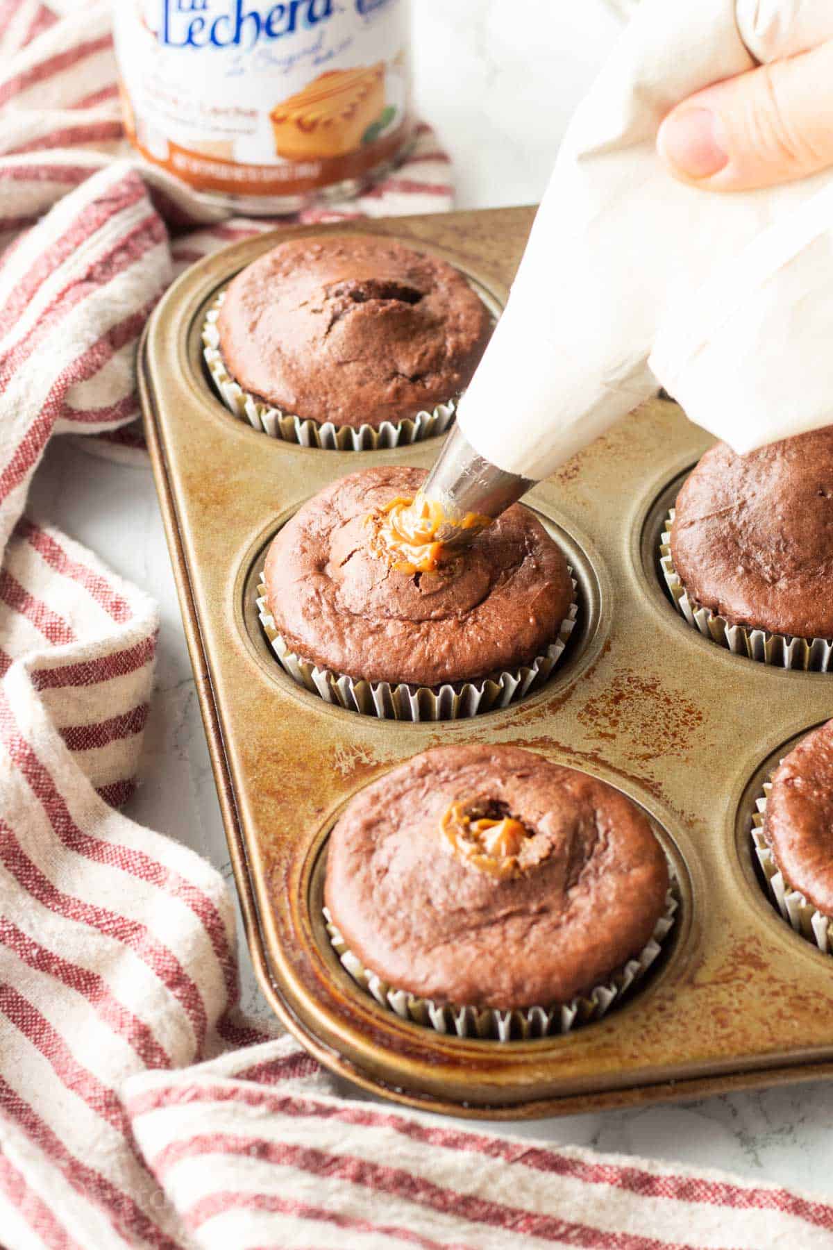 Hand piping dulce de leche into baked chocolate cupcakes in metal pan.