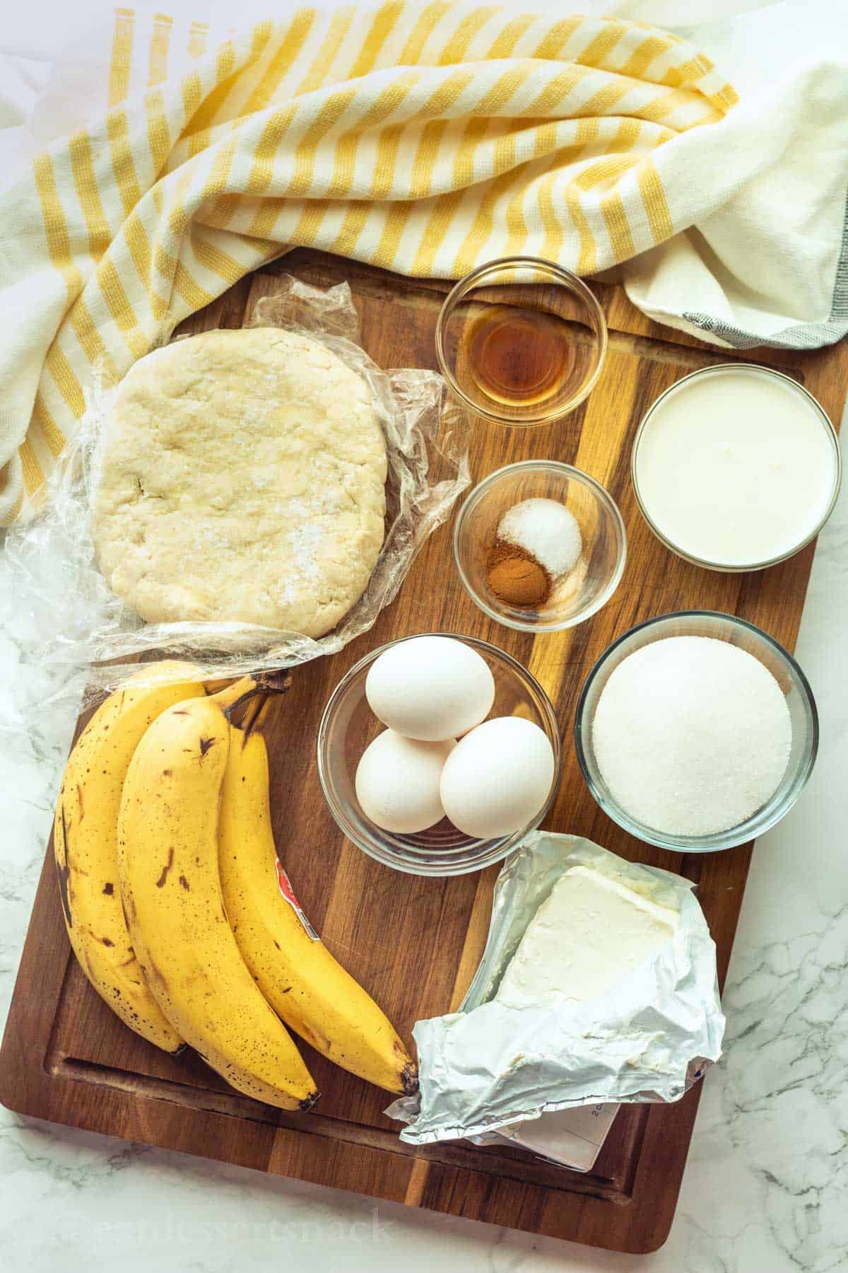 Ingredients for baked banana pie in glass bowls on wood cutting board with yellow striped dish towel.