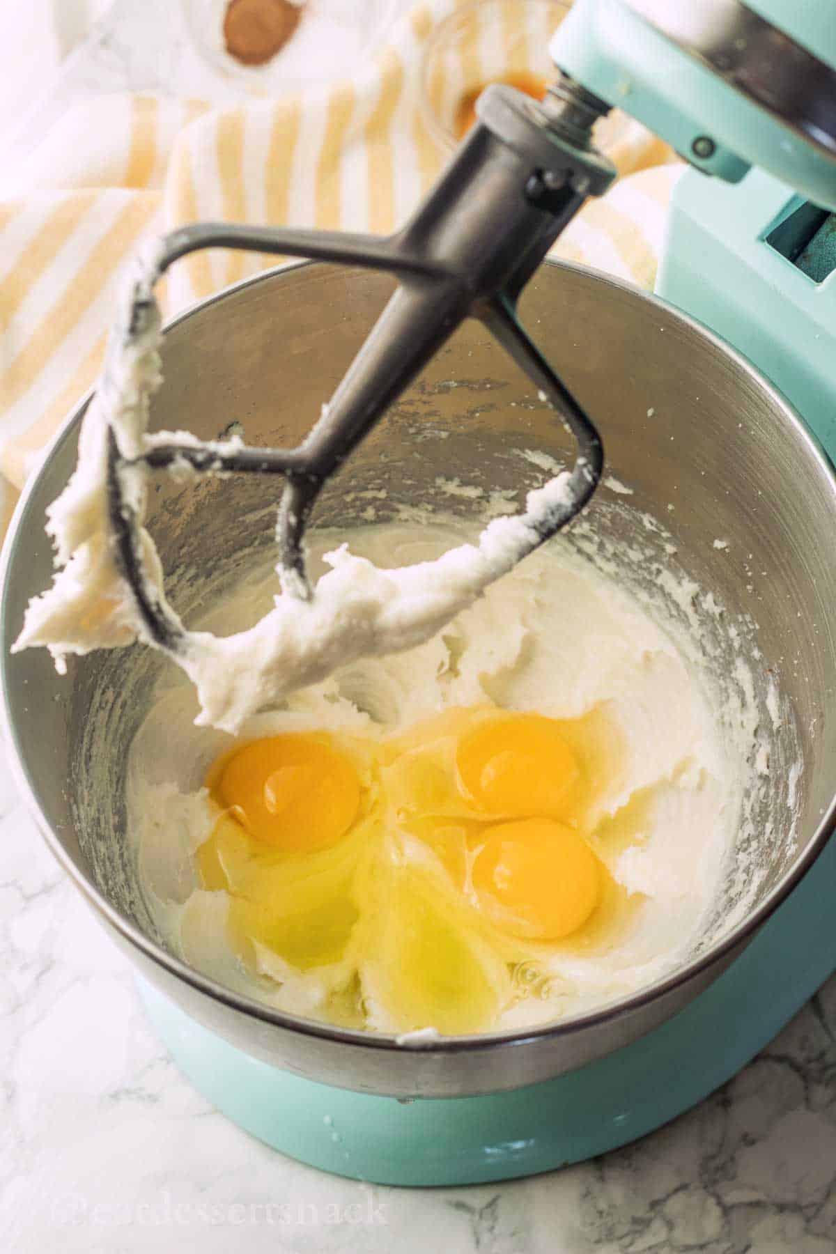 Cream cheese and eggs in mixing bowl with metal mixer paddle above.