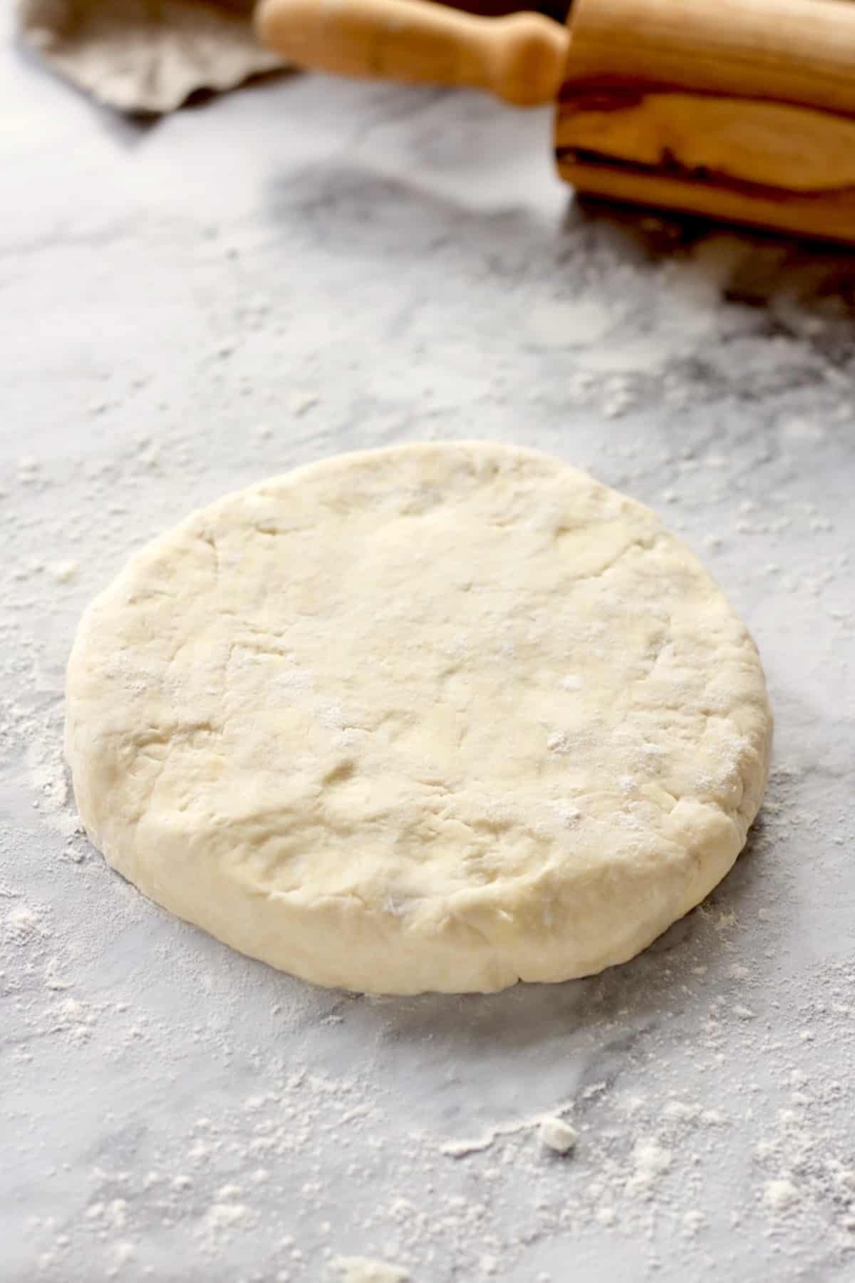 Disc of raw cream cheese pie dough on marble countertop with wooden rolling pin.
