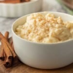 Cooked eggnog rice pudding in white bowl with cinnamon sticks.