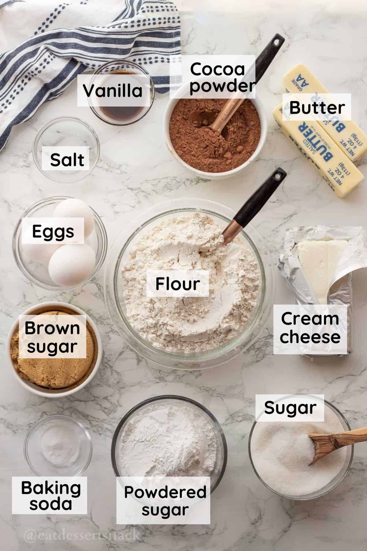 Cocoa powder, butter, vanilla, eggs, salt, cream cheese, sugar, baking soda, brown sugar, and flour in glass bowls on marble countertop.