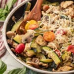 Cooked chicken, zucchini, tomatoes, and mushrooms in metla pan with wooden spoon.