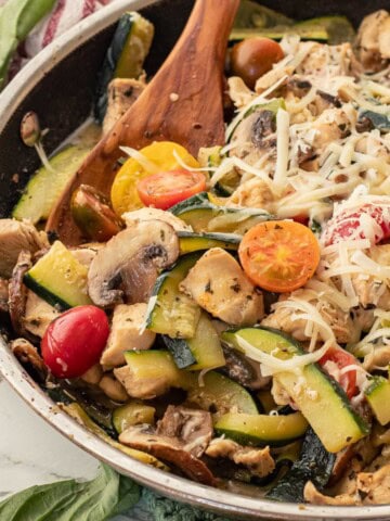 Cooked chicken, zucchini, tomatoes, and mushrooms in metla pan with wooden spoon.