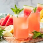 Glass of watermelon mint lemonade on wood cutting board with slice of watermelon and sprig of mint on top.