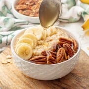 Spoon drizzling honey over white bowl of banana oatmeal.