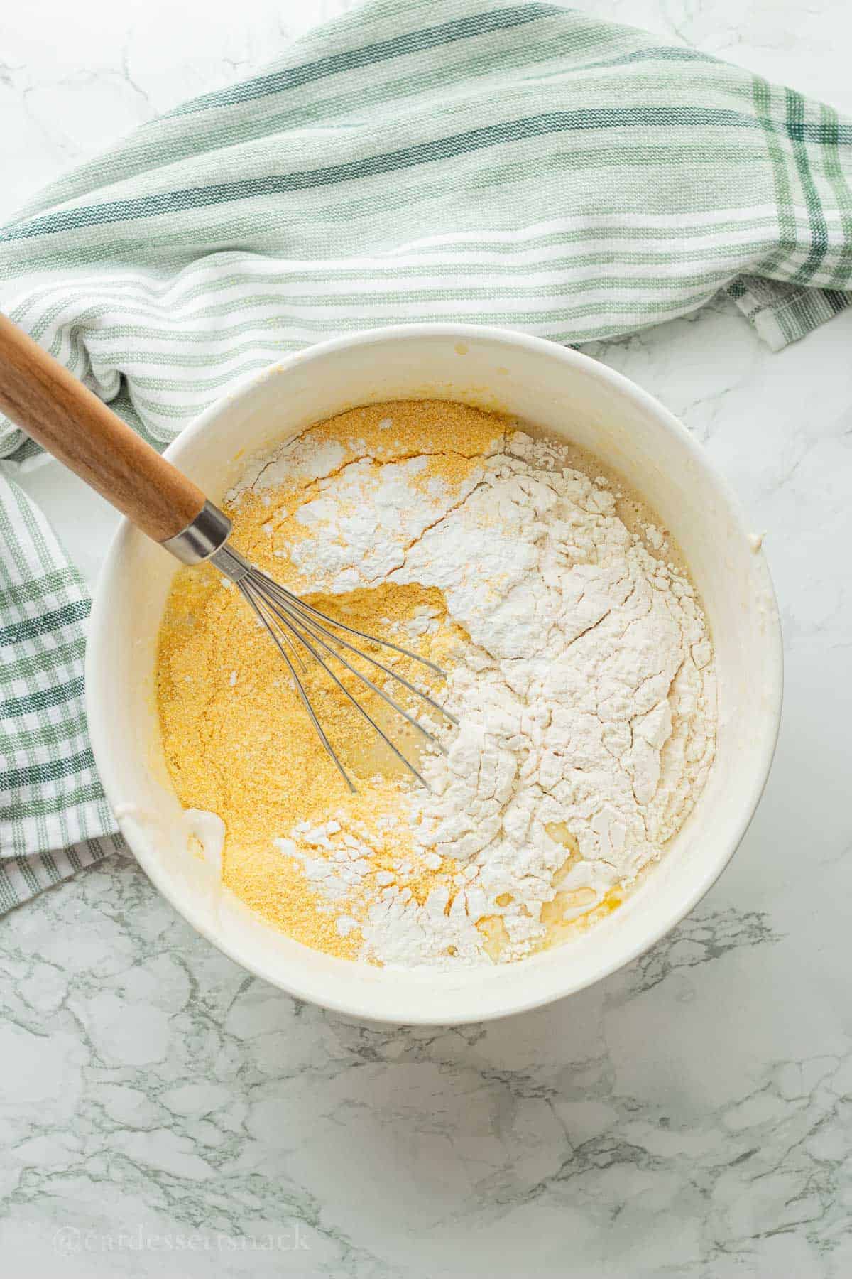 Cornmeal, flour, salt, and baking powder over wet ingredients with whisk in white bowl.