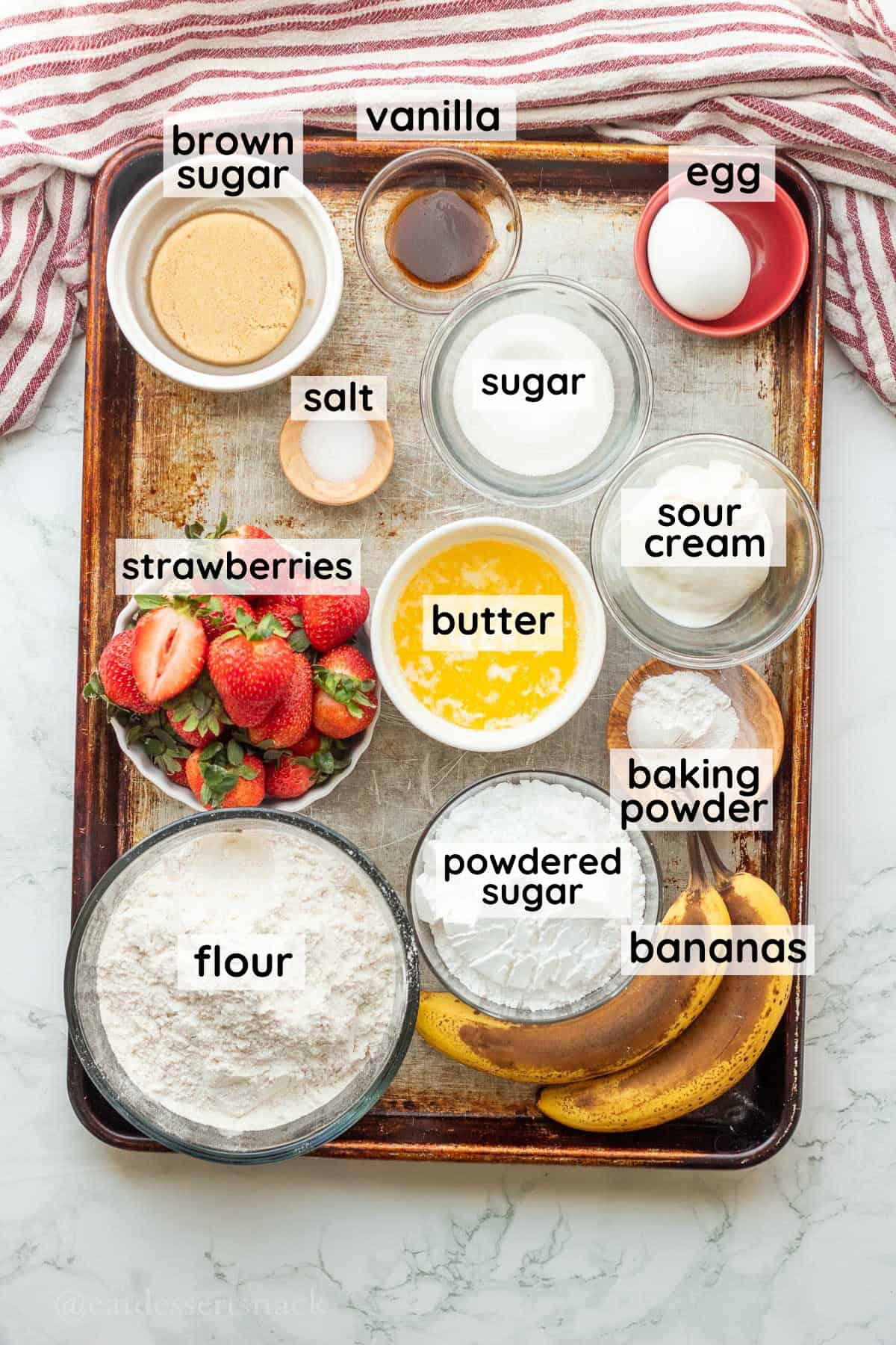 Labeled ingredients for banana strawberry bread in glass bowls on metal pan.