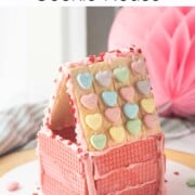 Pink wafer cookie house with Valentine candy on roof on white plate.