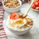 White bowl of yogurt with honey, granola, and strawberry on top with gold spoon.