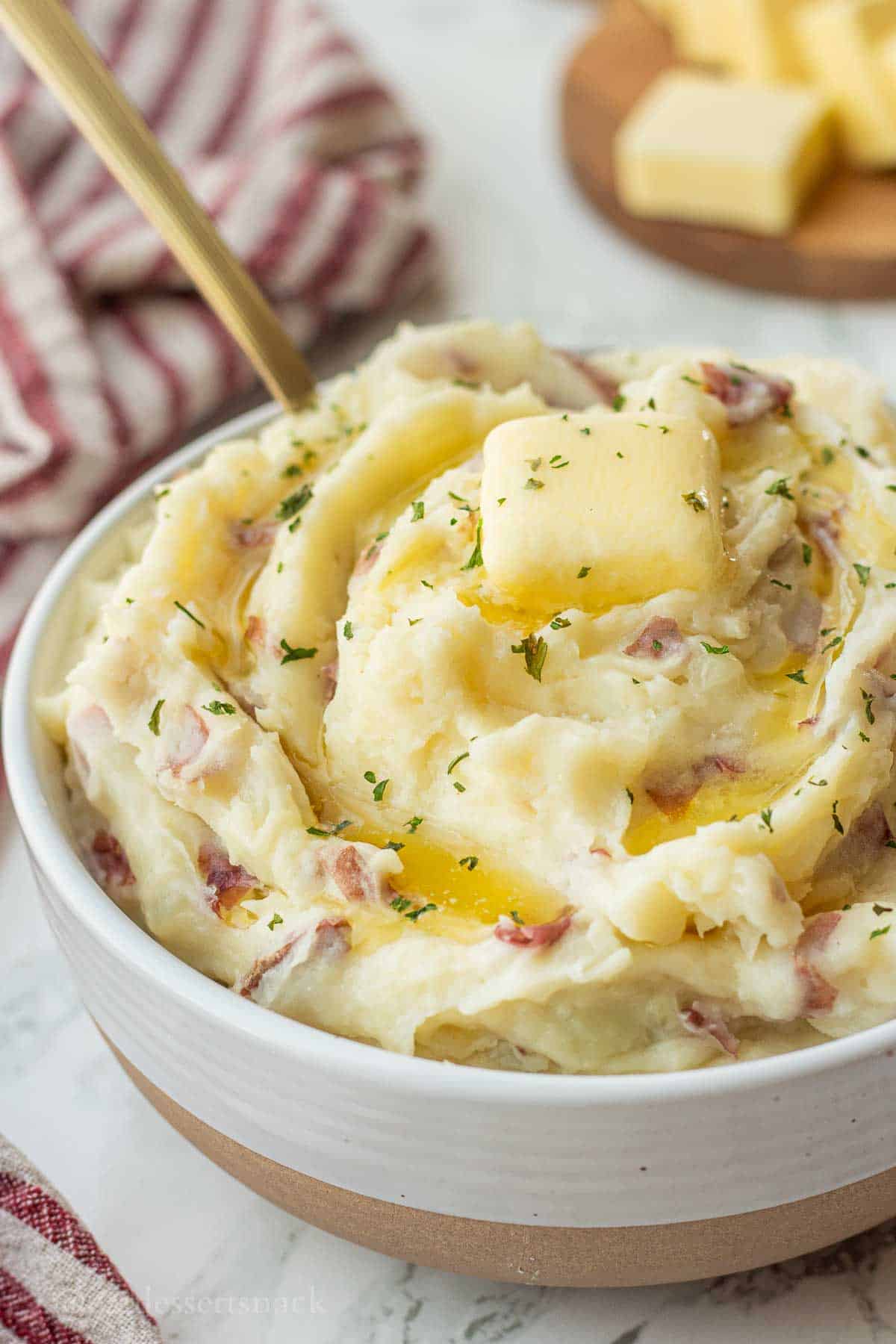 Red skin mashed potatoes with butter on top in white bowl with gold spoon.