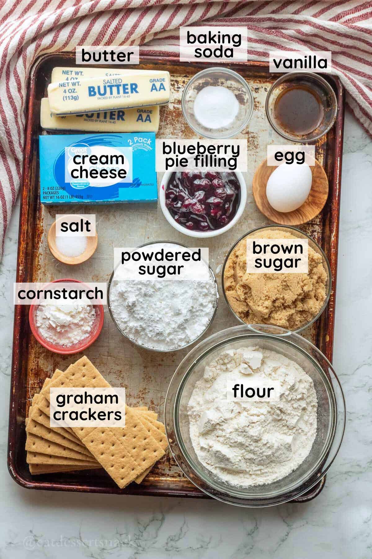 Labeled ingredients for blueberry cheesecake cookies on metal baking pan.