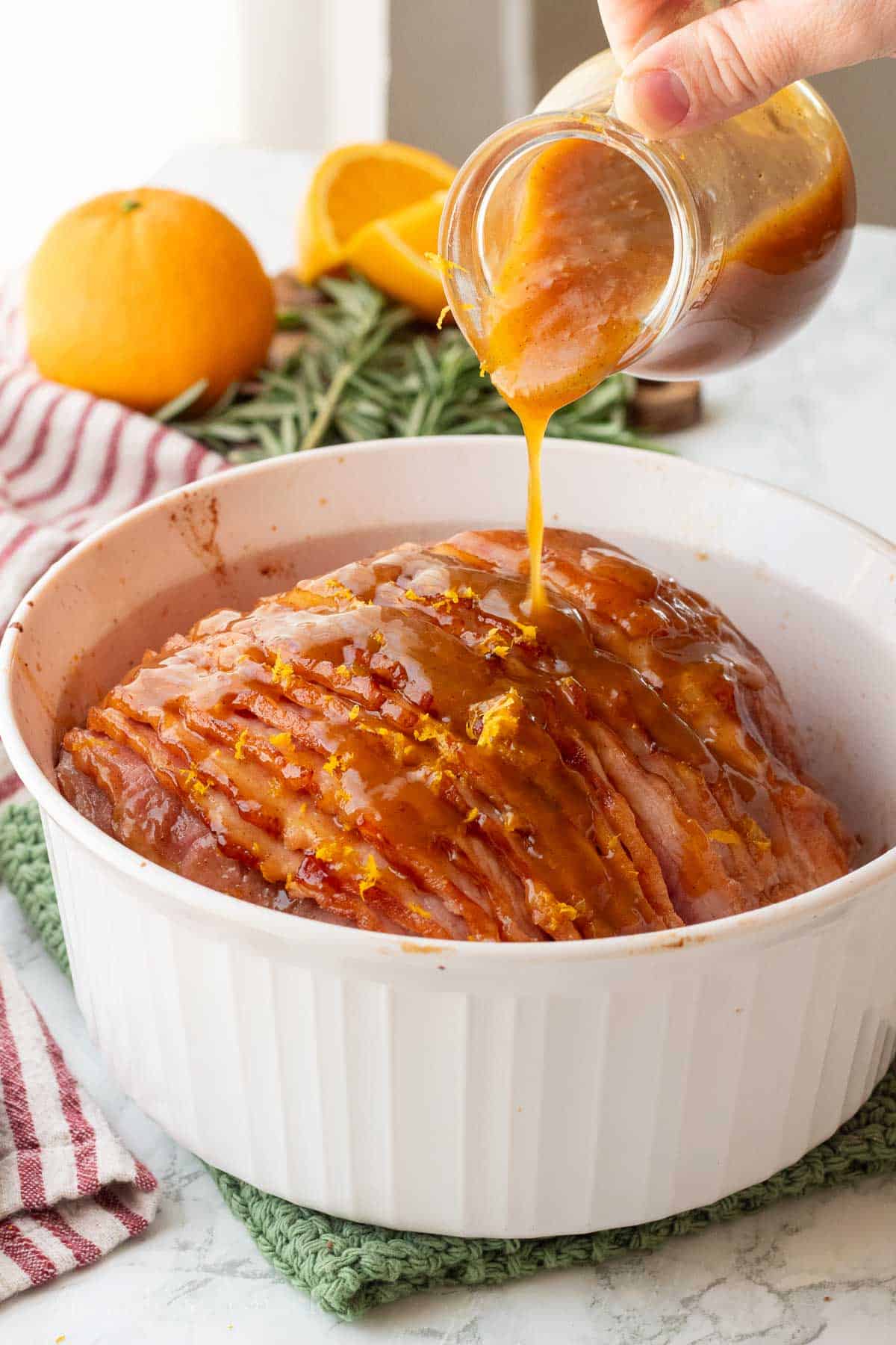 Pouring orange glaze over cooked ham in glass baking dish.