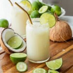 Glass of lime in the coconut drink with straw and lime slice and pitcher of limeade. With coconut shell nearby.