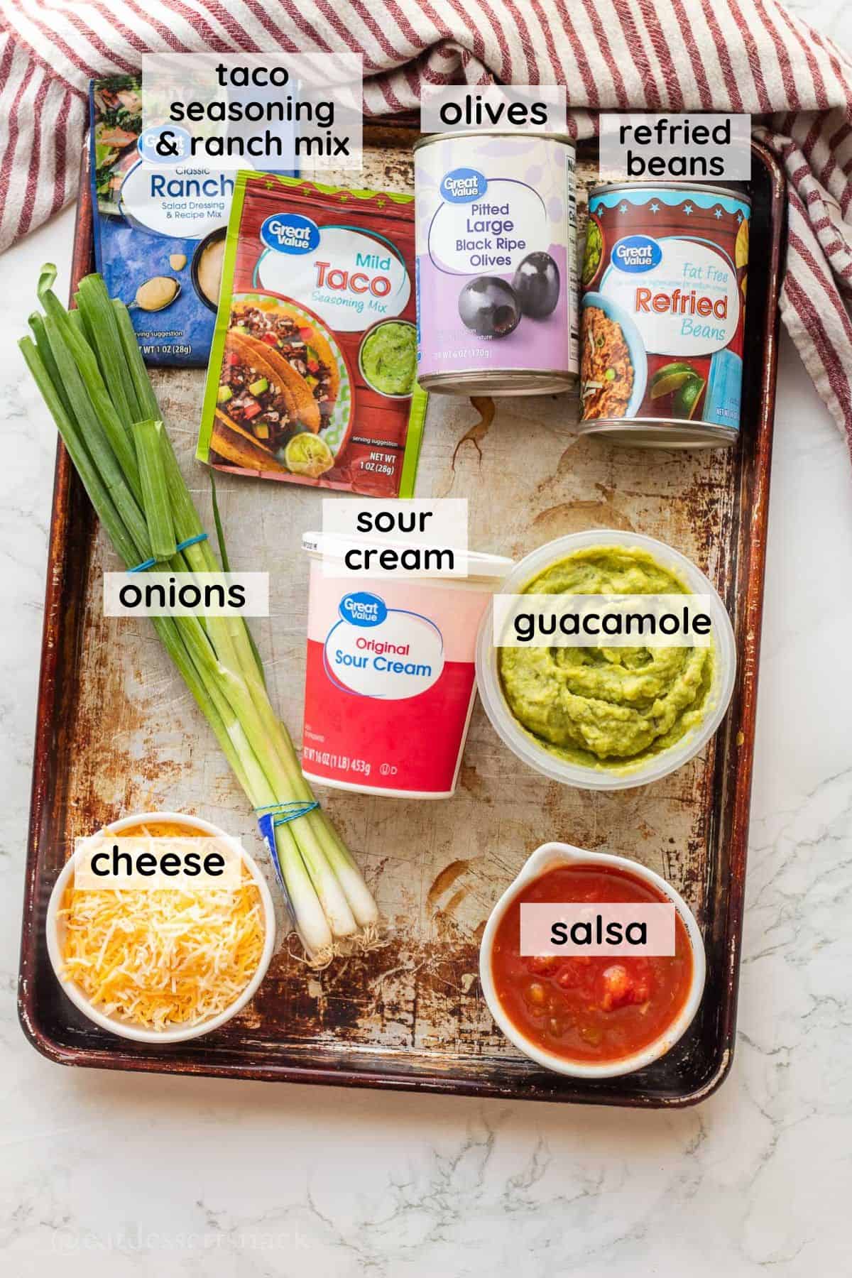 Ingredients for bean dip in glass bowls on metal baking pan. Sour Cream, Guacamole, Salsa, cheese, sour cream, and onions.