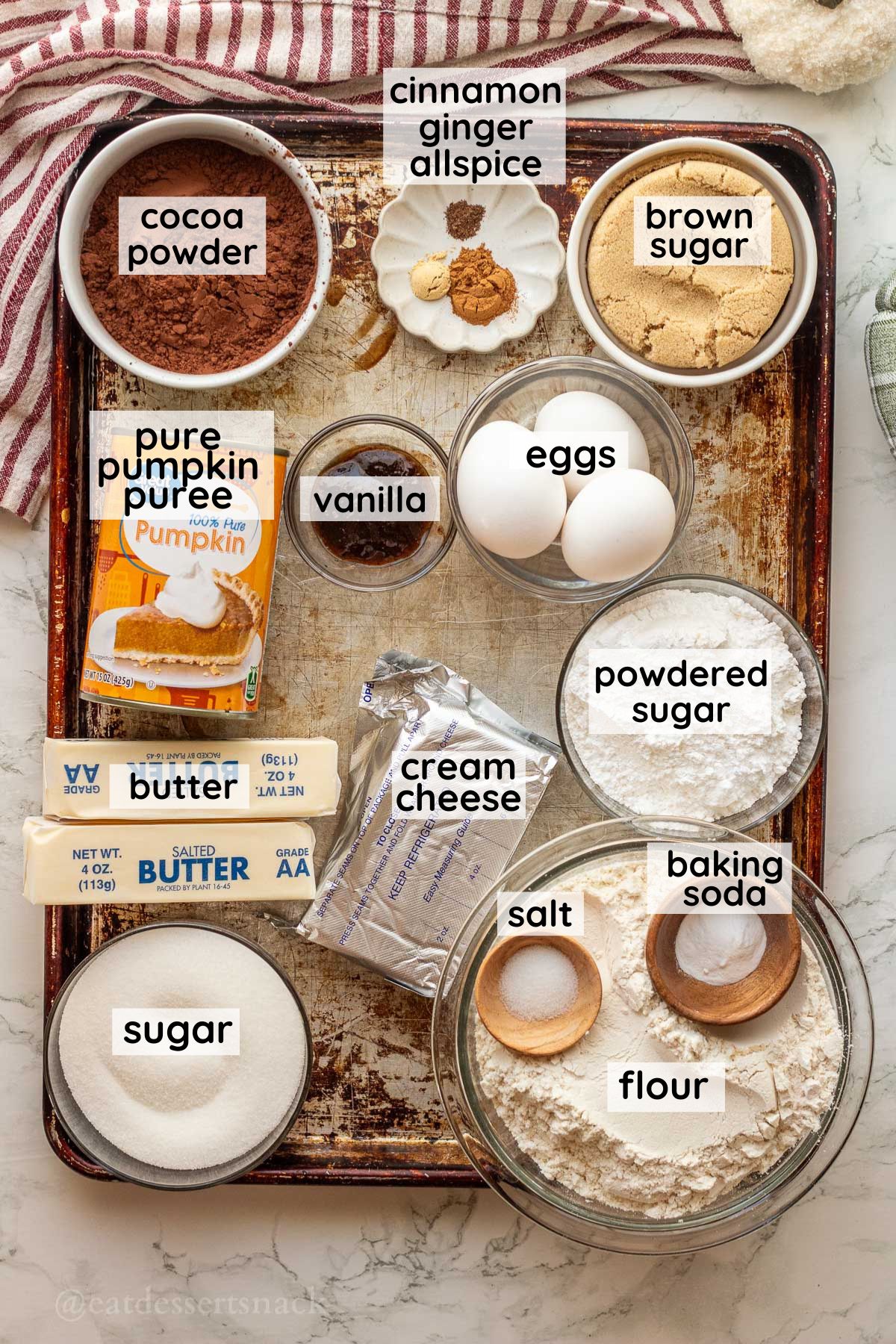 Labeled ingredients for pumpkin cheesecake brownies in glass bowls on metal pan.